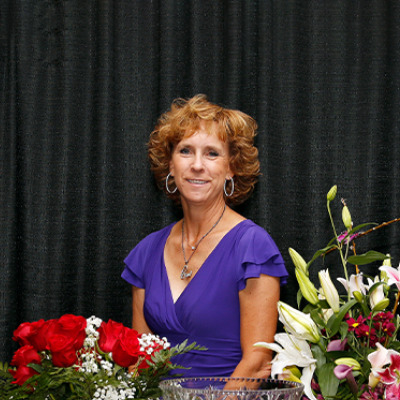 Leslie Lange wearing a purple dress seated behind an arrangement of red roses and white lilies on a table, with a black curtain as the background.