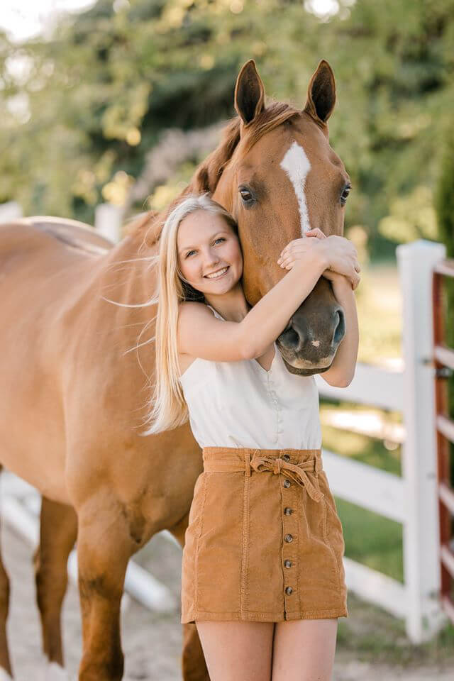 Sydni Loose, a blonde girl smiling while wrapping her arms to hug her horse in a picturesque setting.